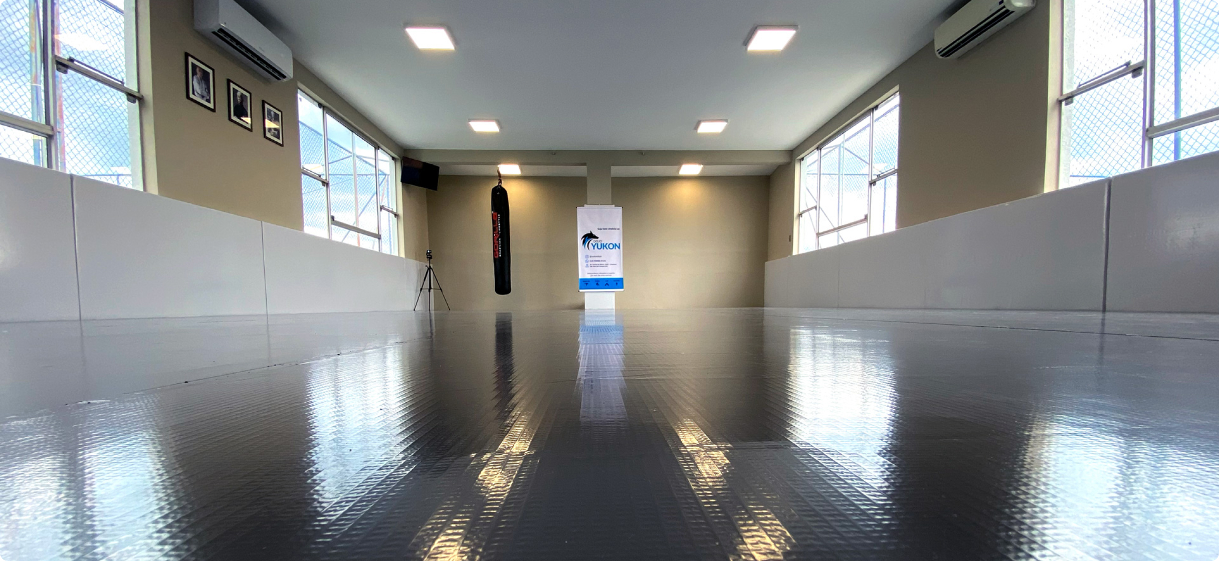Foto do dojo, área de treino de karatê com ar-condicionado, janelas, tv e saco de pancadas